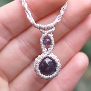 May include: A handmade macrame necklace featuring a dark purple gemstone pendant. The necklace has a light gray cord with a woven design, and a smaller purple bead is set above the pendant. The necklace is held in a hand.