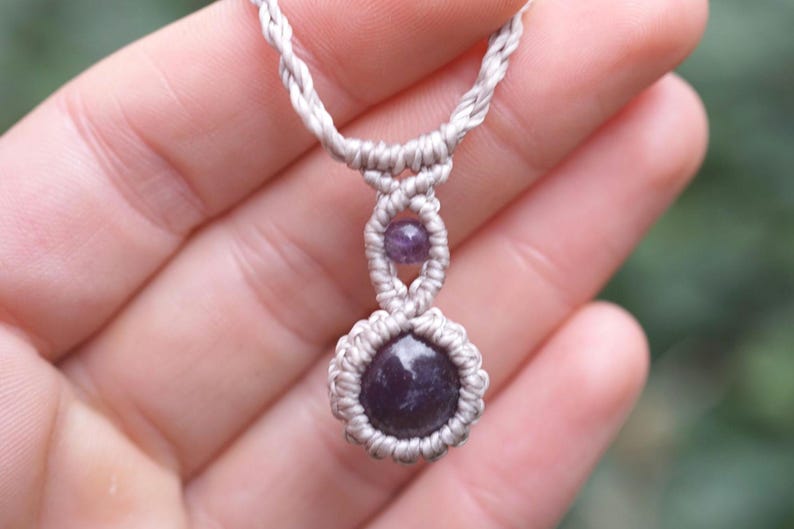 May include: A handmade macrame necklace featuring a dark purple gemstone pendant. The necklace has a light gray cord with a woven design, and a smaller purple bead is set above the pendant. The necklace is held in a hand.