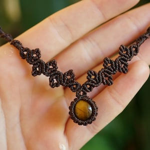 Puede incluir: Un collar de macram&eacute; marr&oacute;n con un colgante central de piedra ojo de tigre circular. El cord&oacute;n est&aacute; intrincadamente anudado en un patr&oacute;n geom&eacute;trico repetitivo que se extiende a lo largo de la cadena, creando un dise&ntilde;o tejido alrededor de la piedra.