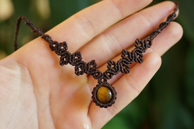 Puede incluir: Un collar de macram&eacute; marr&oacute;n con un colgante central de piedra ojo de tigre circular. El cord&oacute;n est&aacute; intrincadamente anudado en un patr&oacute;n geom&eacute;trico repetitivo que se extiende a lo largo de la cadena, creando un dise&ntilde;o tejido alrededor de la piedra.
