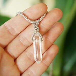 May include: A clear, hexagonal, double-terminated quartz crystal pendant suspended from a light beige, braided macrame cord. The crystal is held in place by a decorative knotting pattern that secures the stone at its center point.