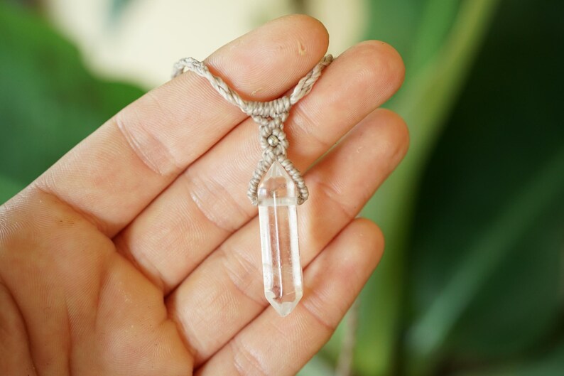 May include: A clear, hexagonal, double-terminated quartz crystal pendant suspended from a light beige, braided macrame cord. The crystal is held in place by a decorative knotting pattern that secures the stone at its center point.