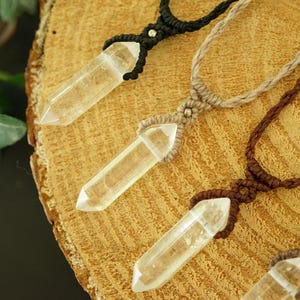 May include: Four necklaces featuring clear, pointed crystal pendants suspended on woven cord in black, beige, and brown. Each pendant is secured with a macrame knot and a small metallic bead, showcasing a natural, geometric design with clear quartz.