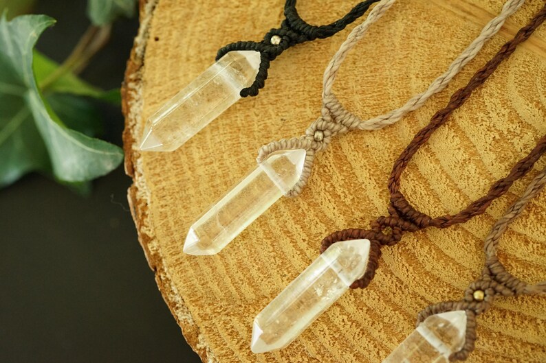 May include: Four necklaces featuring clear, pointed crystal pendants suspended on woven cord in black, beige, and brown. Each pendant is secured with a macrame knot and a small metallic bead, showcasing a natural, geometric design with clear quartz.