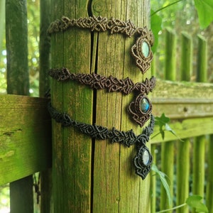 May include: Three macrame necklaces in shades of brown and black are displayed on a wooden post. Each necklace features a central gemstone pendant. The background includes a green fence and foliage, creating a natural setting.