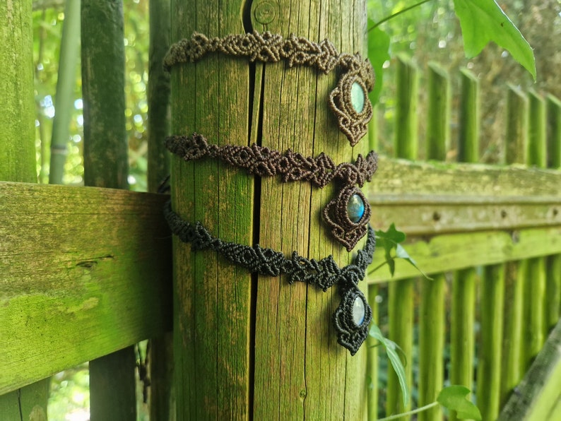 May include: Three macrame necklaces in shades of brown and black are displayed on a wooden post. Each necklace features a central gemstone pendant. The background includes a green fence and foliage, creating a natural setting.