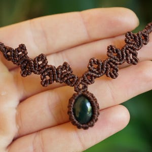 Puede incluir: Un collar de macram&eacute; marr&oacute;n con un colgante central de piedra ovalada de color verde oscuro. El cord&oacute;n est&aacute; intrincadamente anudado en un patr&oacute;n geom&eacute;trico repetitivo que se extiende a lo largo de la cadena, creando un dise&ntilde;o texturizado.