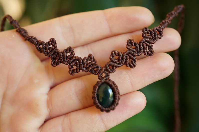Puede incluir: Un collar de macram&eacute; marr&oacute;n con un colgante central de piedra ovalada de color verde oscuro. El cord&oacute;n est&aacute; intrincadamente anudado en un patr&oacute;n geom&eacute;trico repetitivo que se extiende a lo largo de la cadena, creando un dise&ntilde;o texturizado.