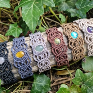May include: Six macrame bracelets with gemstone accents rest on a piece of wood, surrounded by green leaves. The bracelets feature various colors of macrame cord and gemstones, including clear, yellow, green, turquoise, and purple stones.