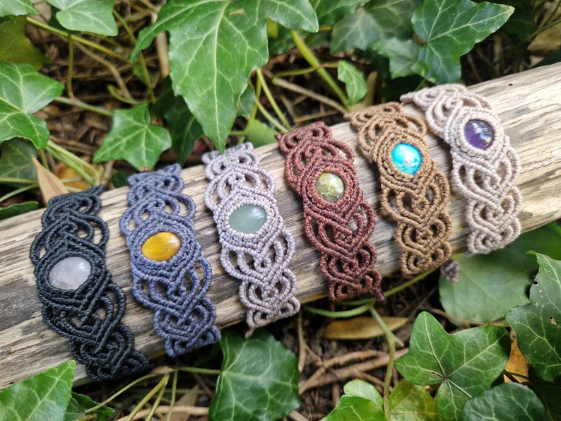 May include: Six macrame bracelets with gemstone accents rest on a piece of wood, surrounded by green leaves. The bracelets feature various colors of macrame cord and gemstones, including clear, yellow, green, turquoise, and purple stones.