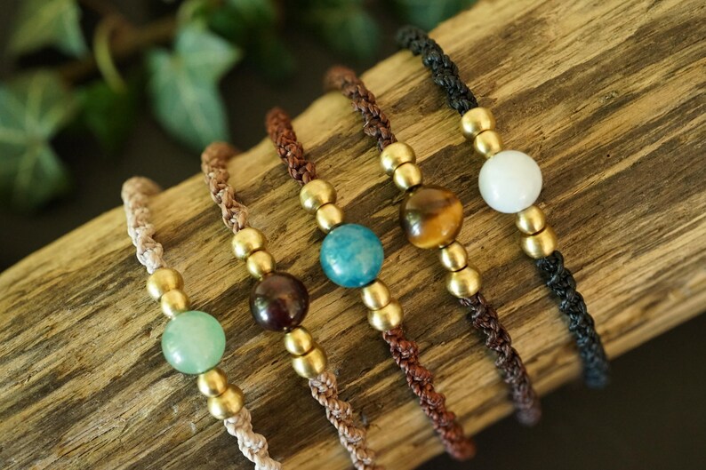 May include: Close-up of five handmade macrame bracelets with gemstone beads. Each bracelet features a different colored gemstone: green, dark red, blue, brown, and white. The bracelets have gold-colored metal beads and are displayed on a piece of wood.