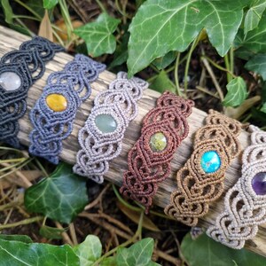 May include: A collection of handmade macrame bracelets in various colors, each featuring a central gemstone. The bracelets are displayed on a piece of wood, surrounded by green foliage. Gemstones include amethyst, turquoise, and yellow stones.