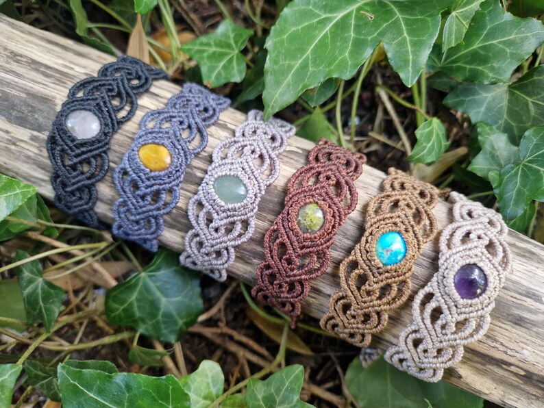 May include: A collection of handmade macrame bracelets in various colors, each featuring a central gemstone. The bracelets are displayed on a piece of wood, surrounded by green foliage. Gemstones include amethyst, turquoise, and yellow stones.