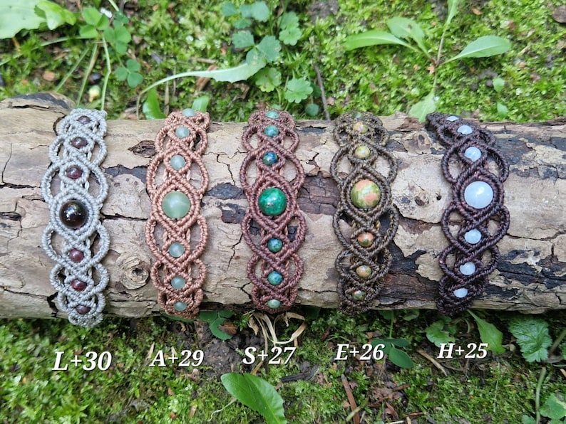 Puede incluir: Cinco pulseras de macram&eacute; con diversas cuentas de piedras preciosas, incluyendo granate, jade y unakita. Las pulseras se exhiben sobre un trozo de madera contra un fondo de follaje verde. Cada pulsera est&aacute; etiquetada con una combinaci&oacute;n de letra y n&uacute;mero.