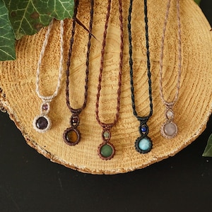 May include: A collection of six macrame necklaces with gemstone pendants. The necklaces feature various colors of cord and gemstones, including amethyst, tiger's eye, green aventurine, turquoise, and rose quartz. They are displayed on a wooden slice.