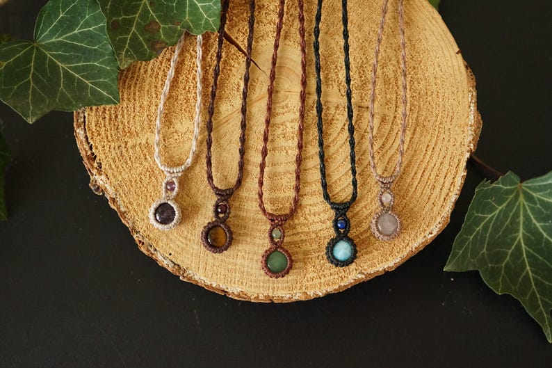 May include: A collection of six macrame necklaces with gemstone pendants. The necklaces feature various colors of cord and gemstones, including amethyst, tiger's eye, green aventurine, turquoise, and rose quartz. They are displayed on a wooden slice.