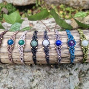May include: A collection of handmade macrame bracelets, each featuring a different gemstone. The bracelets are in various colors, including brown, blue, green, and purple, and are adorned with small gold beads. They are displayed on a piece of wood.