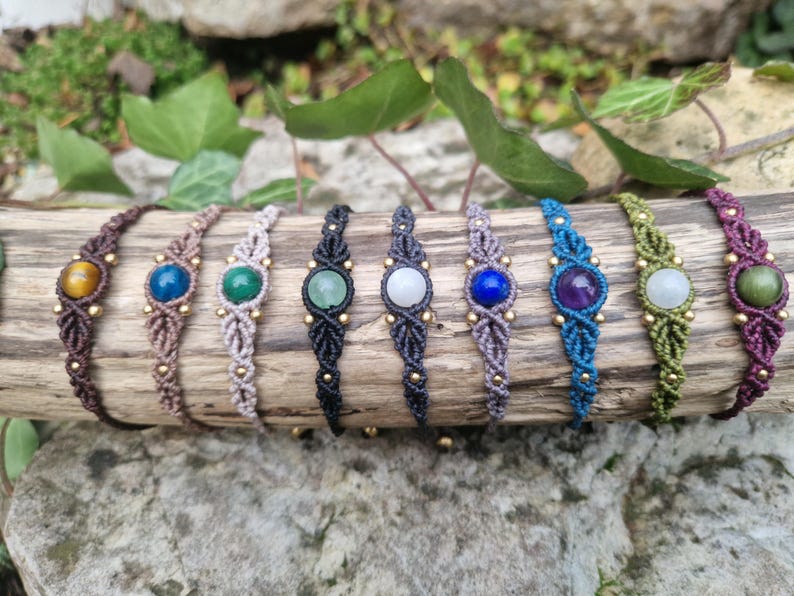 May include: A collection of handmade macrame bracelets, each featuring a different gemstone. The bracelets are in various colors, including brown, blue, green, and purple, and are adorned with small gold beads. They are displayed on a piece of wood.