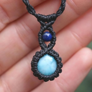 May include: A handmade macrame necklace featuring a round, light blue gemstone pendant. The pendant is surrounded by black macrame knots, with a smaller blue bead above it. The necklace is displayed against a blurred background.