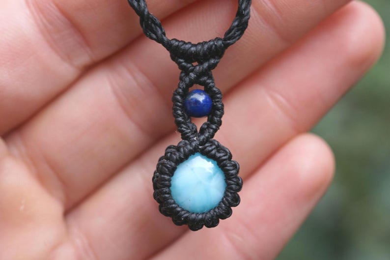 May include: A handmade macrame necklace featuring a round, light blue gemstone pendant. The pendant is surrounded by black macrame knots, with a smaller blue bead above it. The necklace is displayed against a blurred background.