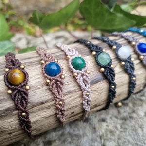 May include: A collection of macrame bracelets with gemstone accents. Each bracelet features a different colored gemstone, including yellow, blue, green, and white, set in a woven macrame design. The bracelets are adorned with small gold beads.