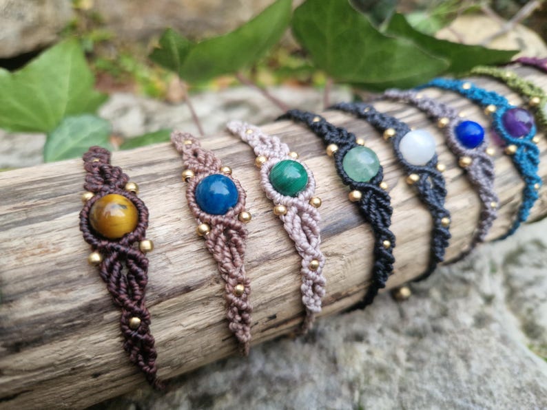 May include: A collection of macrame bracelets with gemstone accents. Each bracelet features a different colored gemstone, including yellow, blue, green, and white, set in a woven macrame design. The bracelets are adorned with small gold beads.