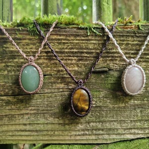 Peut inclure: Trois colliers en macramé faits à la main avec des pendentifs ovales en pierres précieuses. Les pierres sont vertes, marron et roses, chacune sertie dans un cadre en macramé tissé. Les colliers sont exposés sur une surface en bois vieilli.