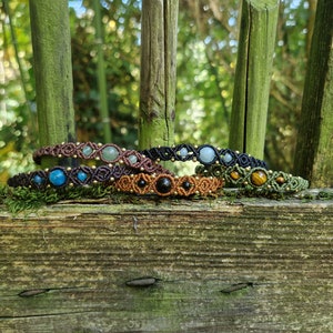 May include: A collection of macrame bracelets with various gemstones, including blue, green, and black stones, displayed on a weathered wooden surface. The bracelets are in shades of brown, black, and green, with gold-colored accents.
