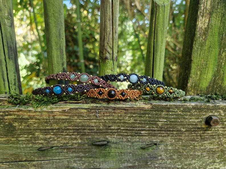 May include: A collection of macrame bracelets with various gemstones, including blue, green, and black stones, displayed on a weathered wooden surface. The bracelets are in shades of brown, black, and green, with gold-colored accents.