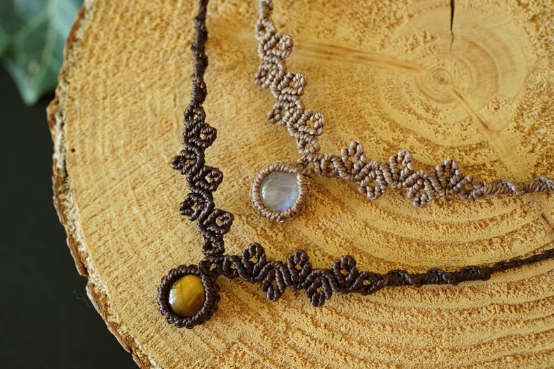 Puede incluir: Dos collares de macram&eacute; con intrincados dise&ntilde;os de cord&oacute;n anudado. Un collar es de color marr&oacute;n oscuro con una piedra de ojo de tigre redonda, y el otro es de color marr&oacute;n claro con un colgante de cabuj&oacute;n redondo, transl&uacute;cido y azul p&aacute;lido.