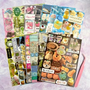 May include: A collection of colorful sticker sheets featuring various designs, including food, animals, and Japanese text. The stickers showcase illustrations of desserts, drinks, and other cute objects. A pink tweezer is also visible.