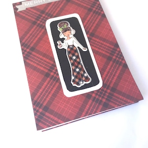May include: A red and black plaid card with a silver banner that says "Merry Christmas". A cartoon illustration of a woman in a plaid skirt and white shirt is on the card.