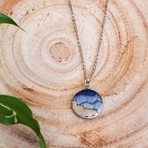 May include: A silver necklace with a round pendant featuring a landscape design in shades of blue and beige. The pendant is suspended from a delicate chain. The necklace is displayed on a wooden surface with green foliage.