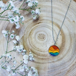 May include: A silver-toned necklace with a circular pendant featuring a colorful, embroidered landscape design. The pendant showcases a gradient of orange, yellow, blue, and green. The necklace rests on a wooden slice, with white flowers in the background.