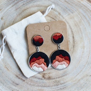 May include: Embroidered dangle earrings featuring a mountain range design in shades of red, orange, and white on a black background. The earrings are displayed on a brown card with a small white drawstring bag.