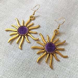 以下が含まれることがあります： 中央に紫色の織り模様が入った、金色のサンバーストイヤリングのペア。 イヤリングはフック留めで、質感のある明るい色の表面に表示されています。 サンバーストのデザインは細長い光線が特徴です。