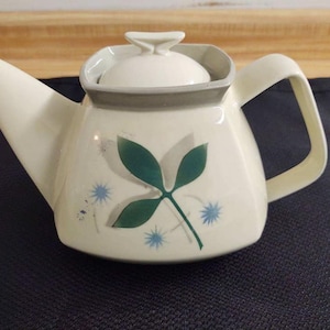 May include: A white ceramic teapot with a gray lid and a green leaf design. The teapot has a square body and a curved spout. The design features green leaves and blue flowers.