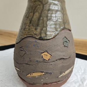 May include: A handmade ceramic vase with a textured, brown surface. The top section is a darker, glossy brown. The middle section features carved fish and geometric shapes. The base is a reddish-brown color. A unique, decorative piece.