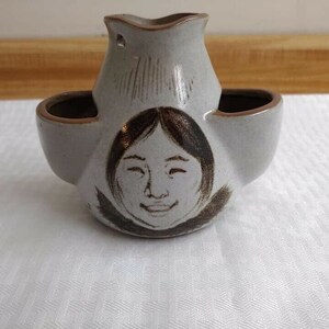 May include: A white ceramic pitcher with a brown face design and two side spouts. The pitcher has a hole in the top for pouring.