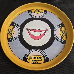 May include: A round, yellow tray with a colorful design. The center features a smiling mouth with red lips and white teeth, surrounded by a black circle. The tray has a light blue background and the name "peter max" is printed on the bottom.