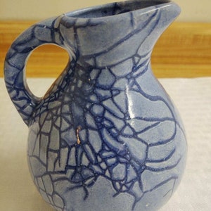 May include: A small, blue ceramic pitcher with a crackled glaze. The pitcher has a round body and a handle.