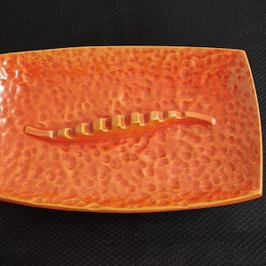 May include: Orange ceramic ashtray with a textured surface and a built-in cigarette rest.