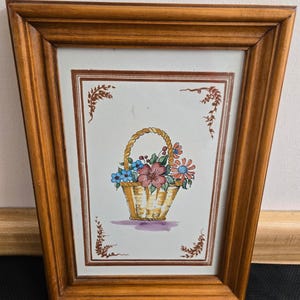 May include: A framed floral print featuring a woven basket overflowing with colorful flowers. The artwork is set against a white background with decorative brown accents and framed in a wooden frame.