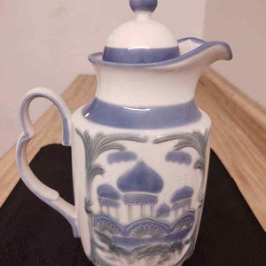 May include: A white and blue ceramic pitcher with a lid. The pitcher features a detailed design of blue domes and foliage. The handle has a heart-shaped design. The pitcher is likely for serving beverages.