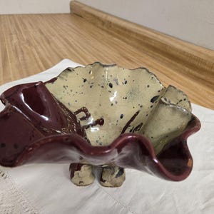 May include: A ceramic bowl with a ruffled edge, featuring a two-tone design. The outer rim is a deep burgundy, while the interior has a speckled beige finish with dark blue accents. The bowl rests on three small, rounded feet.