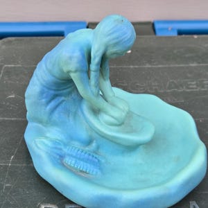 May include: A matte turquoise and blue figurine depicting a person kneeling while grinding grain with a stone tool on a flat base. The figure has long hair and is positioned over a circular basin. An ear of corn is molded onto the base surface.
