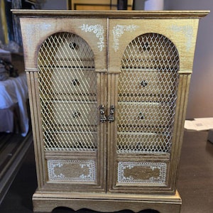 May include: A gold-painted jewelry armoire with two doors, each featuring a wire mesh screen and decorative white floral accents. The armoire has multiple drawers and a decorative top and base.