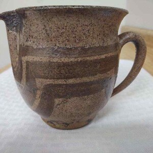 May include: A brown and white ceramic pitcher with a handle. The pitcher has a textured surface and a geometric design.