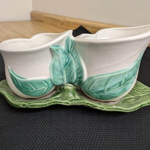 May include: A white ceramic planter with green leaf details, shaped like two calla lilies, sits on a green leaf-shaped base.