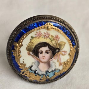 May include: A round, antique trinket box with a portrait of a woman wearing a hat. The portrait is framed in gold and set against a white background, surrounded by a blue enamel border. The box has a silver-colored metal rim.